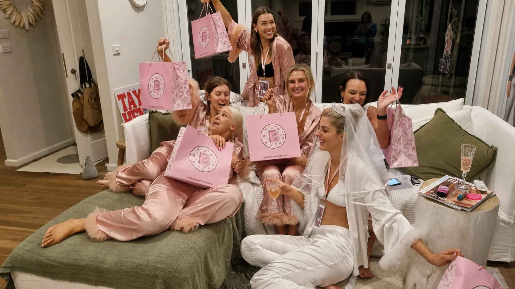 Hens party guests in blush satin pyjamas and a bride in white cheering with pink StayBliss gift bags in a styled living room